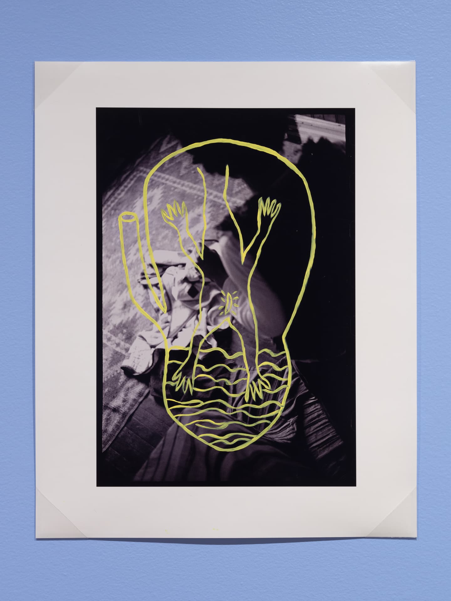 A black and white photograph features a distorted outline of a figure with multiple arms drawn in neon yellow. The figure is placed over an abstract background that includes a textured surface and draped fabric. The image is mounted on a white mat and displayed against a light blue wall.