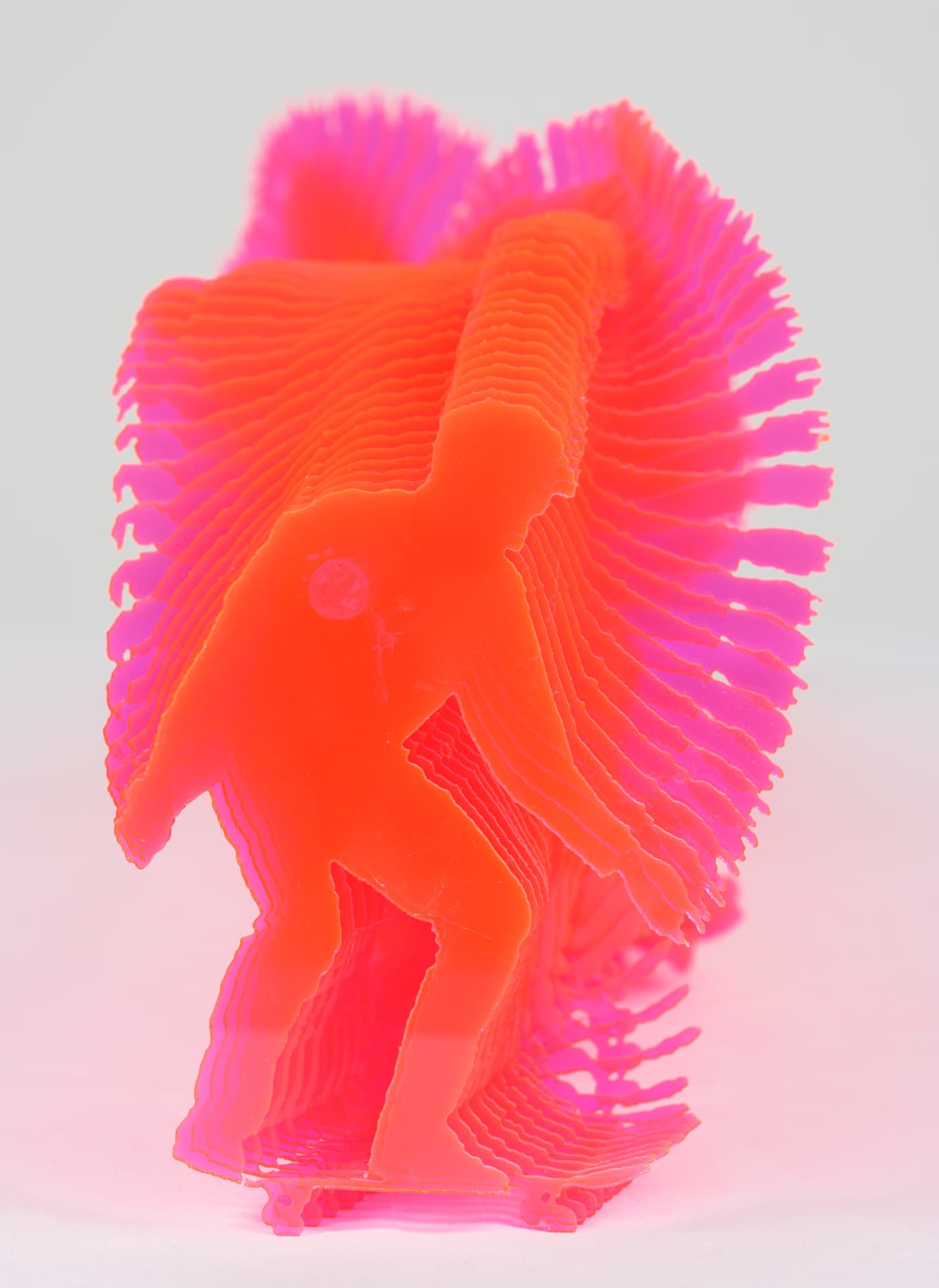A layered, translucent sculpture showcasing a figure in a dynamic pose, appearing to skate or glide. The sculpture is primarily bright neon orange with accents of pink, creating a vibrant and eye-catching effect. The layers are intricately detailed, allowing light to pass through and highlighting the contours of the figure against a soft, neutral background.