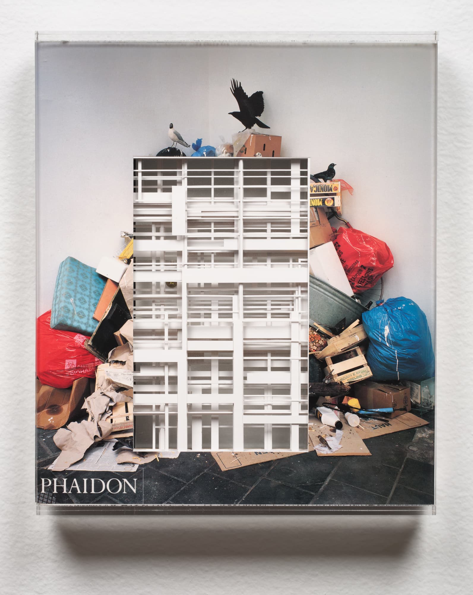 A book cover featuring a collage of contrasting elements: a white architectural model of a building is overlaid on a chaotic scene of litter and discarded items, including colorful trash bags and cardboard boxes. Two birds are perched above the mess, and the title "PHAIDON" is displayed at the bottom. The background is a neutral wall.