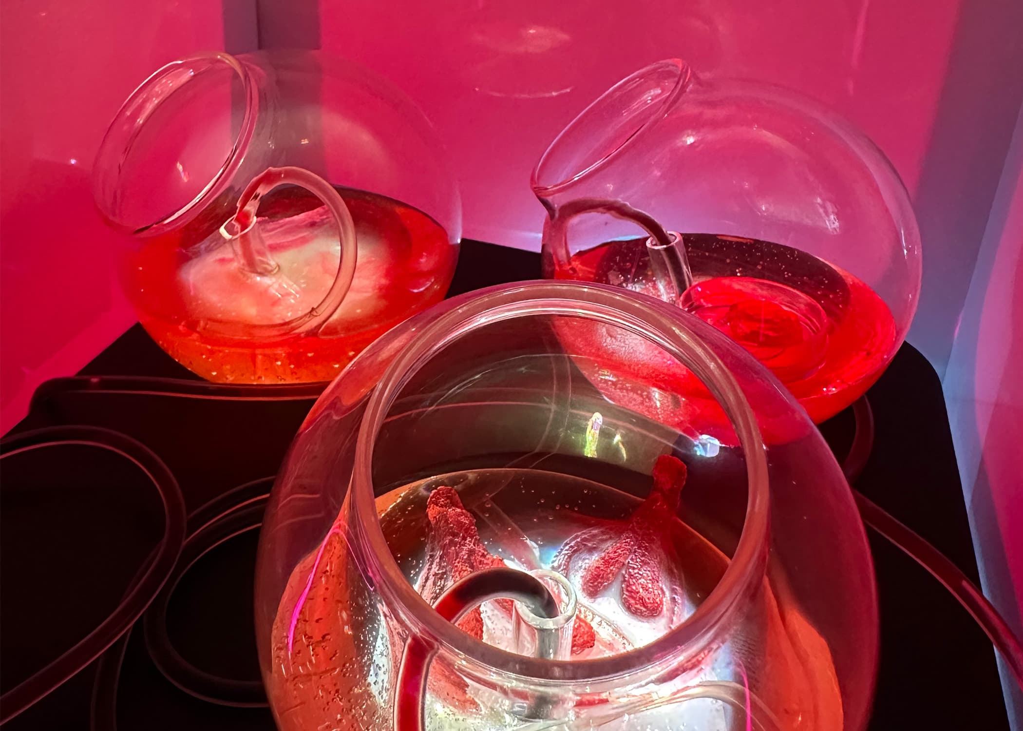 Three glass bowls filled with colorful liquids are arranged on a black surface. The bowls are slightly tilted, revealing one with an orange liquid, one with a red liquid, and another with a clear liquid that has hints of red. A tube connects to each bowl, and vibrant colored lighting, primarily in pink, creates a glowing effect around them.
