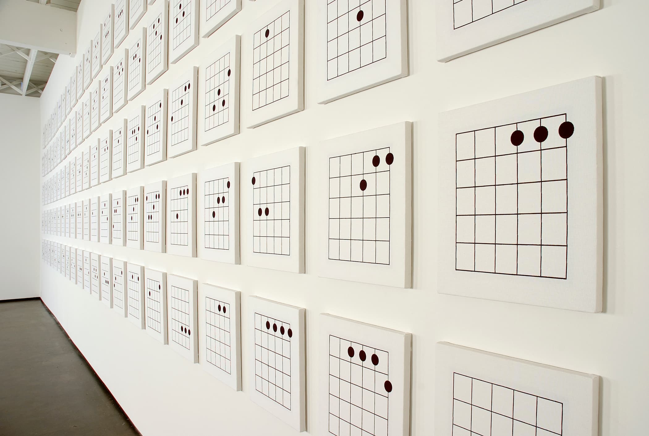 A wall is covered with a grid of framed artworks, each featuring a diagram of guitar chords. The diagrams consist of a series of horizontal lines and dots, arranged in a uniform pattern across a bright white background. The frames are simple and light-colored, and the overall presentation emphasizes a minimalist aesthetic.