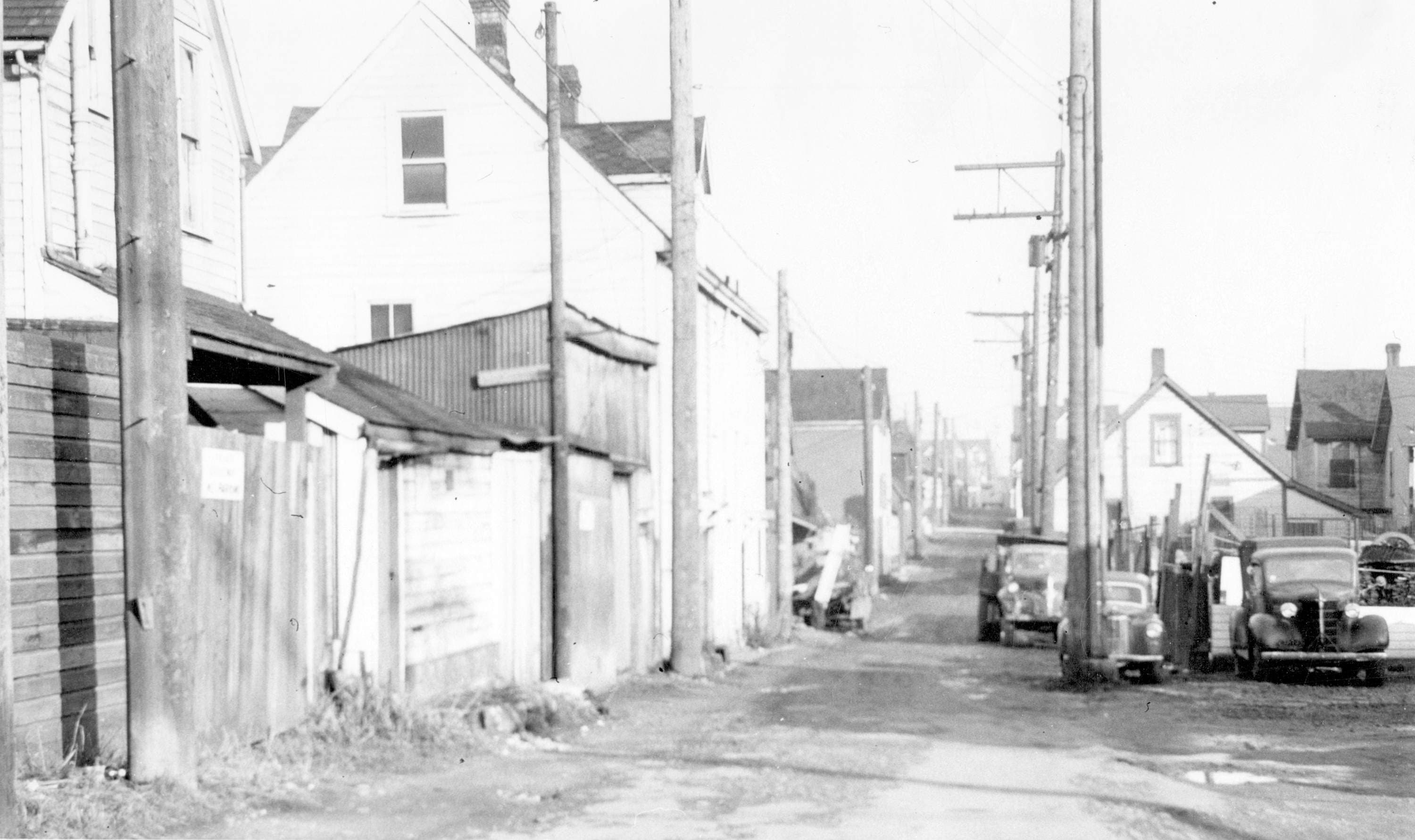 A black and white image of a narrow dirt alley lined with wooden buildings and power poles. On the left side, there are simple structures, some with signs, and on the right side, two old vehicles are parked near a fence. The background features more houses with sloped roofs, and power lines run along the alley, creating a sense of depth. The scene appears to be from an earlier era, evoking a nostalgic atmosphere.