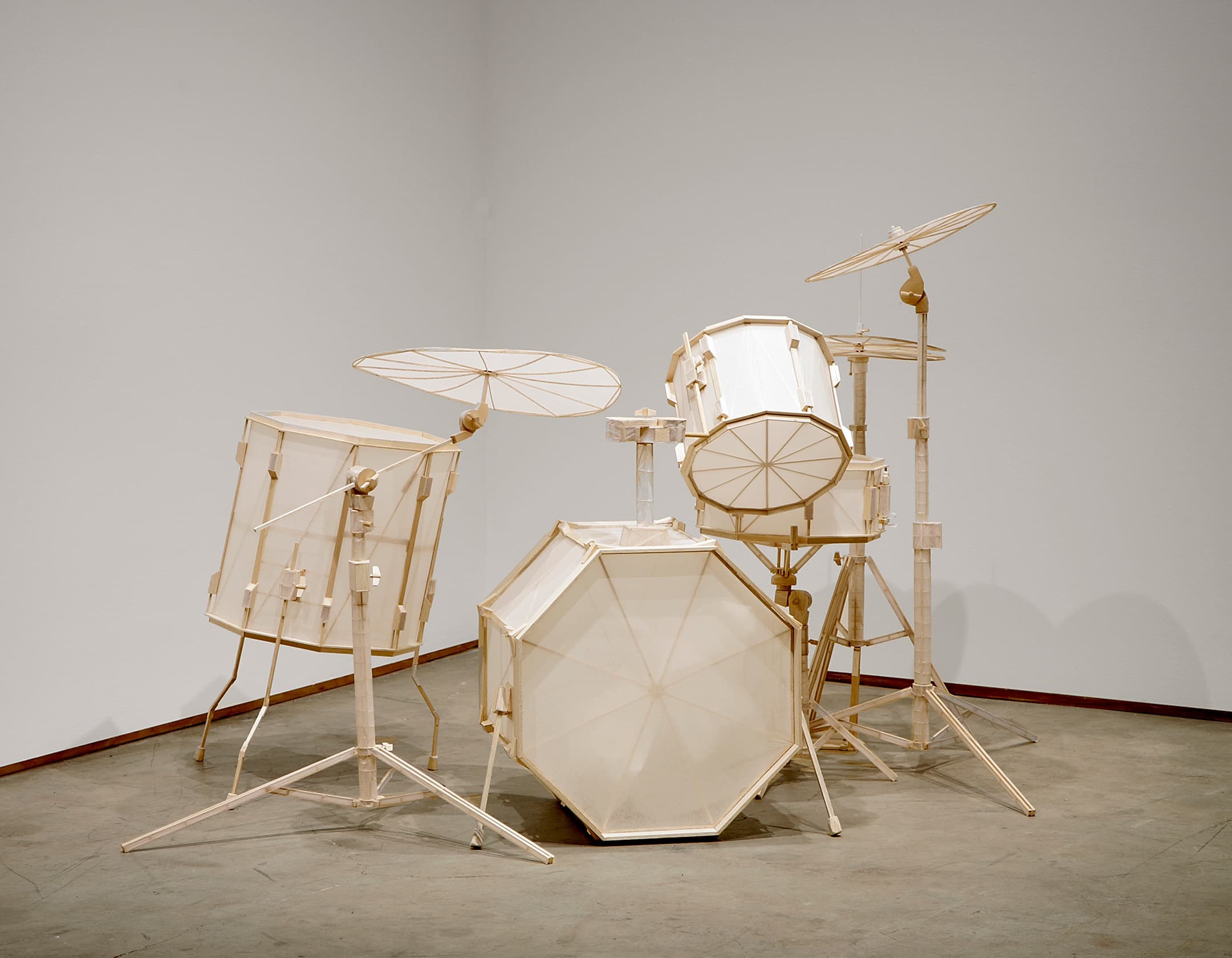 A minimalist drum set made from light-colored materials, featuring a bass drum, snare drum, and cymbals, all with geometric designs and a sculptural appearance. The drums are mounted on thin, simple stands, set against a plain white wall and grey floor.