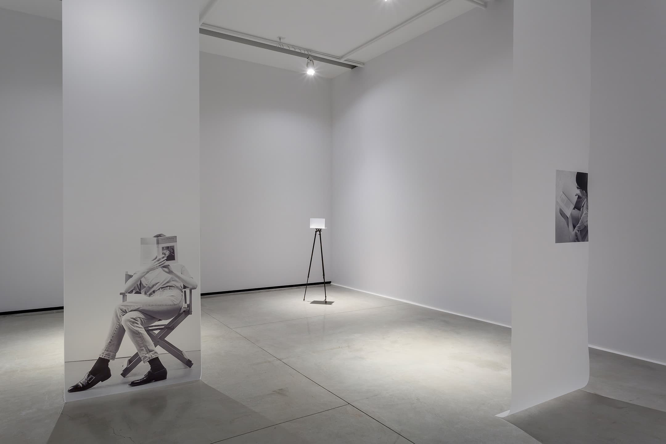 A sparse gallery space with white walls and a concrete floor. A large black-and-white photograph of a seated figure reading is displayed on one wall. In the center, a minimalist lamp on a tripod stand provides soft illumination. A second smaller photograph is mounted on another wall, showing a close-up of a person reading. The lighting creates subtle shadows across the floor.