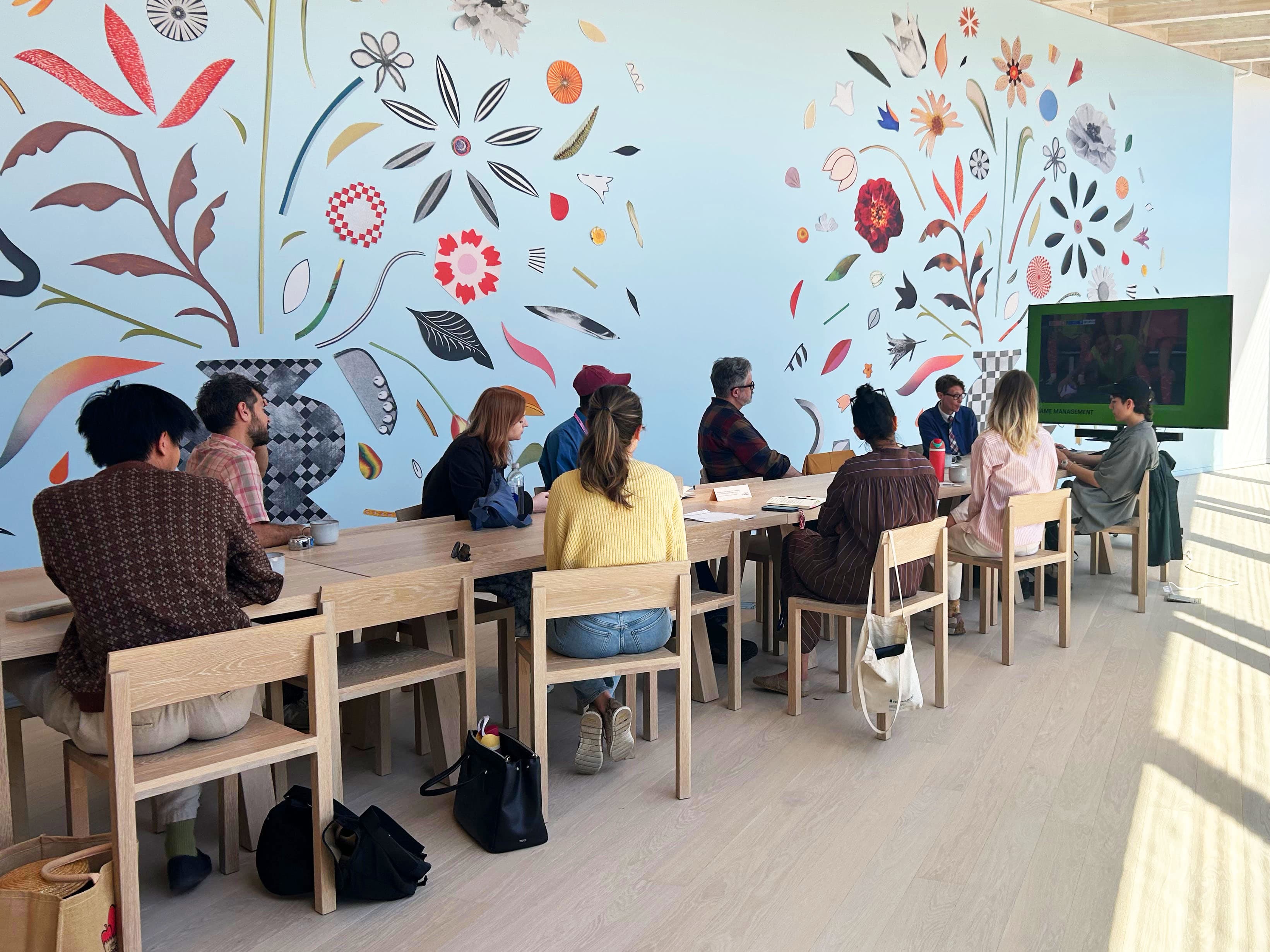 A diverse group of individuals sit at a long wooden table in a brightly lit gallery. They are facing a large screen displaying content related to a presentation. Behind them is a colorful mural featuring various floral and abstract designs against a light blue wall. The participants appear engaged in discussion, with some taking notes. The setting has a modern, open feel with large windows allowing natural light to enter.