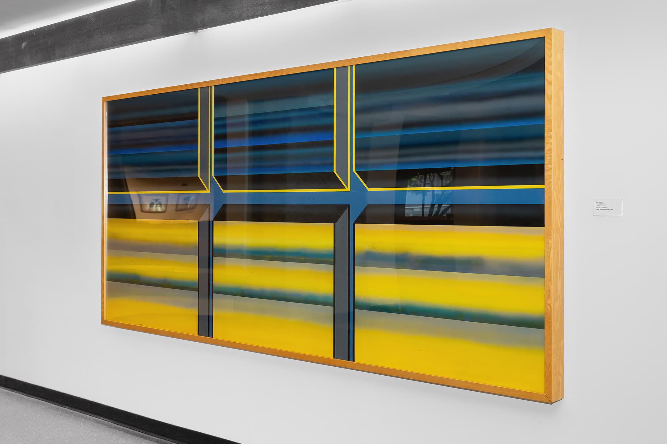 A large abstract painting mounted on a white wall, featuring horizontal bands of blue and yellow, intersected by geometric lines. The artwork has a wooden frame and is illuminated, adding depth to the colors. A small label with information about the artwork is visible to the right.
