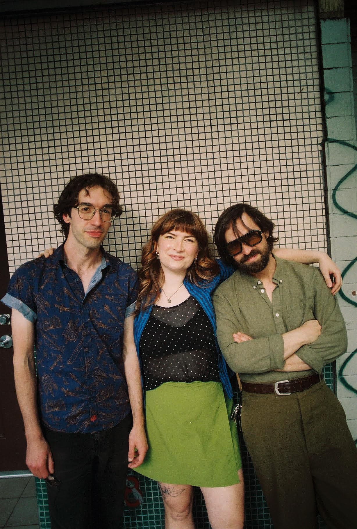 Three individuals pose for a photo against a tiled wall. On the left is a man with curly hair wearing glasses and a blue patterned shirt. In the center is a woman with wavy hair, wearing a black polka dot top and a green skirt, smiling at the camera. On the right is a man with a beard, wearing dark sunglasses and a green shirt, with his arms crossed.