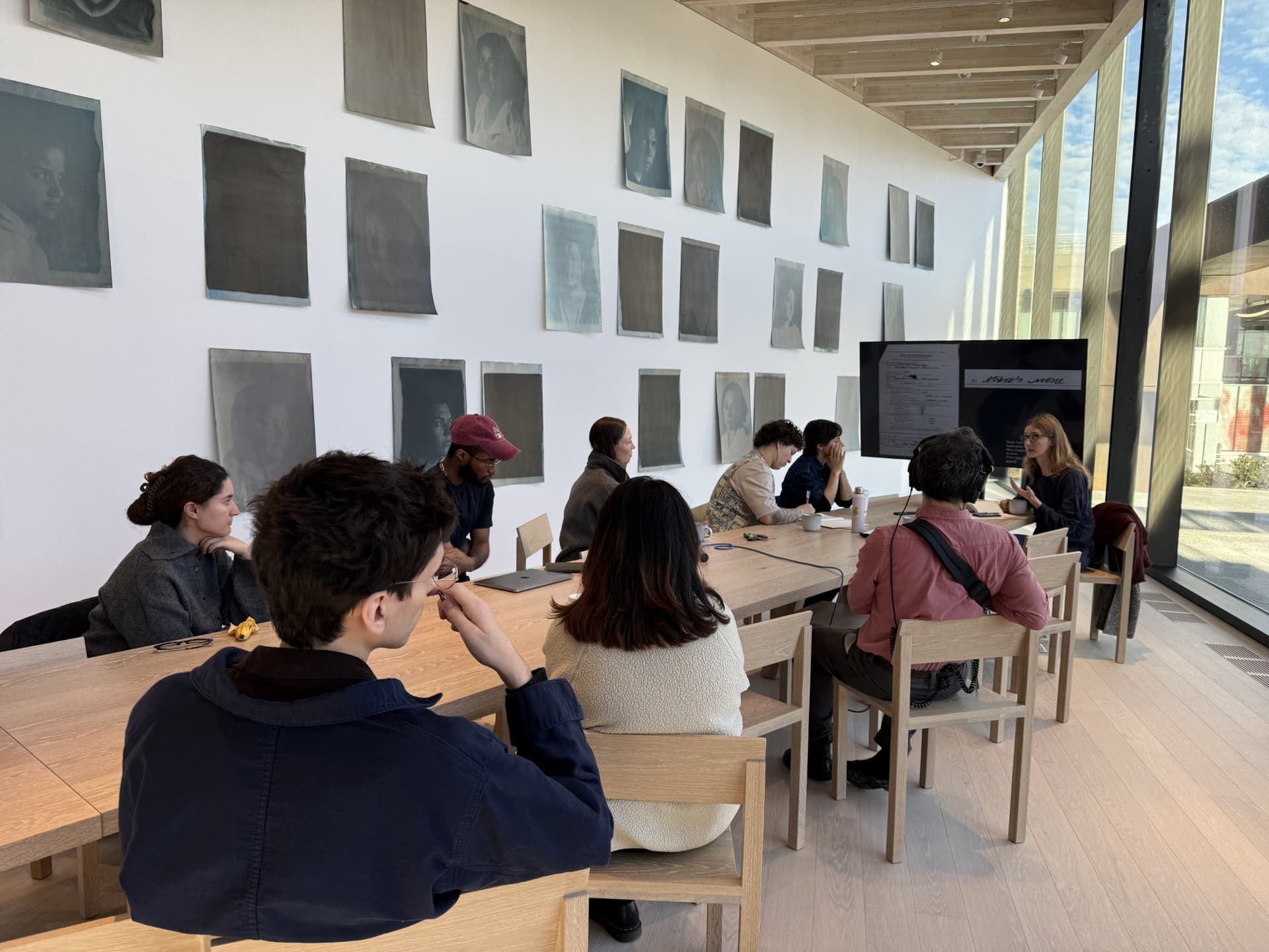 A group of individuals is seated around a wooden table in a well-lit room with large windows. The walls are adorned with various monochromatic portraits. One person is speaking at the front, while others listen attentively; some are taking notes on their laptops. The scene conveys a collaborative meeting or discussion atmosphere.